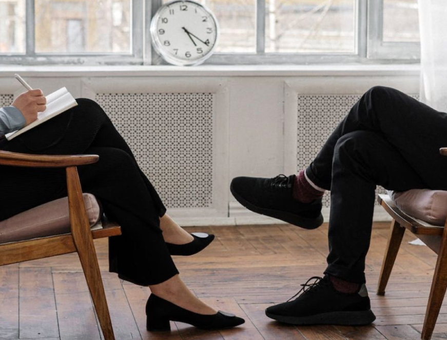 Two people sitting and talking in a room with a window, a clock shows 3:14, one person taking notes, the other with crossed legs, wooden floors, and white curtains.