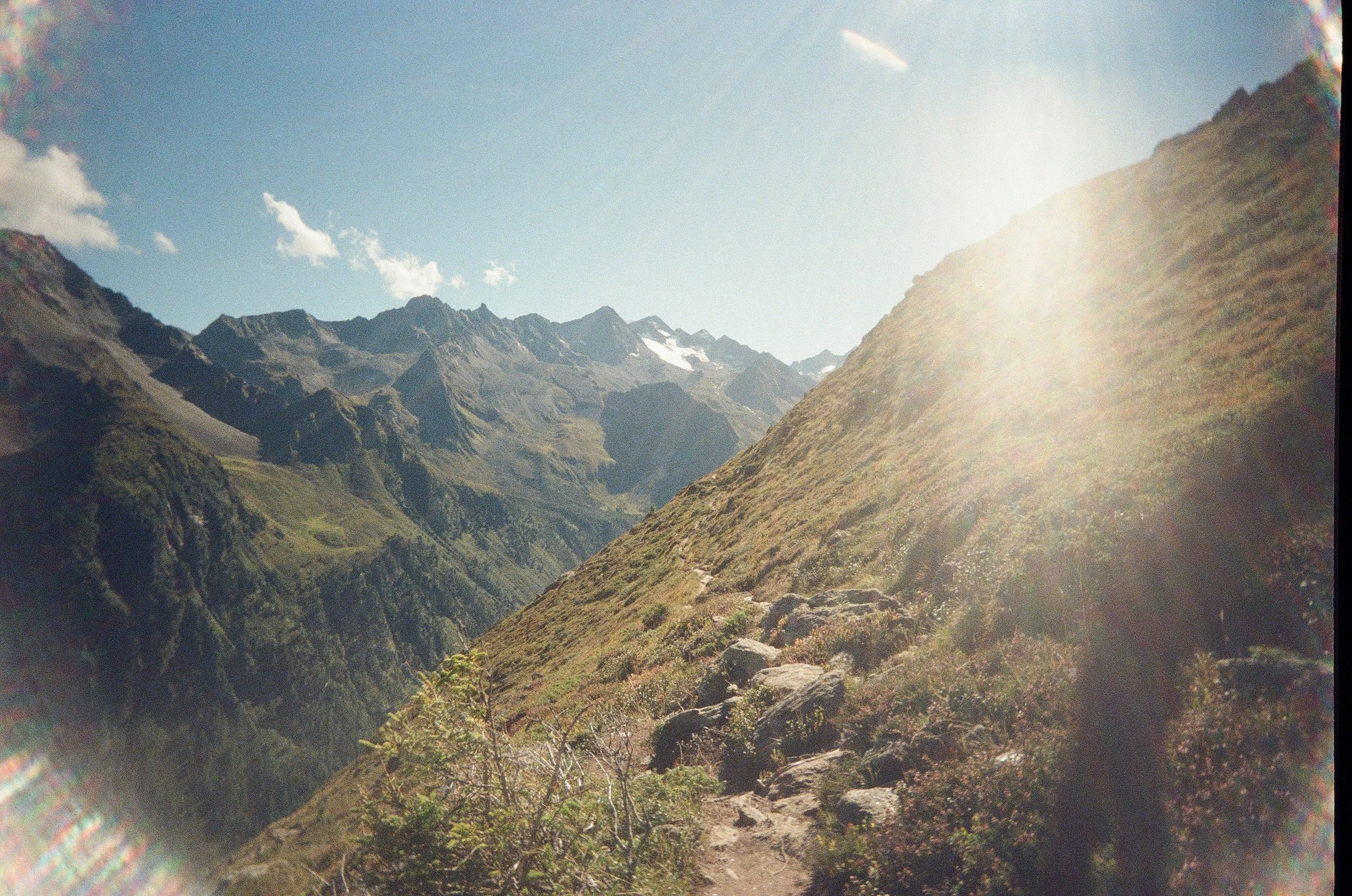 Mountain landscape with rugged peaks, grassy slopes, and a clear sky with some clouds and a visible jet trail.