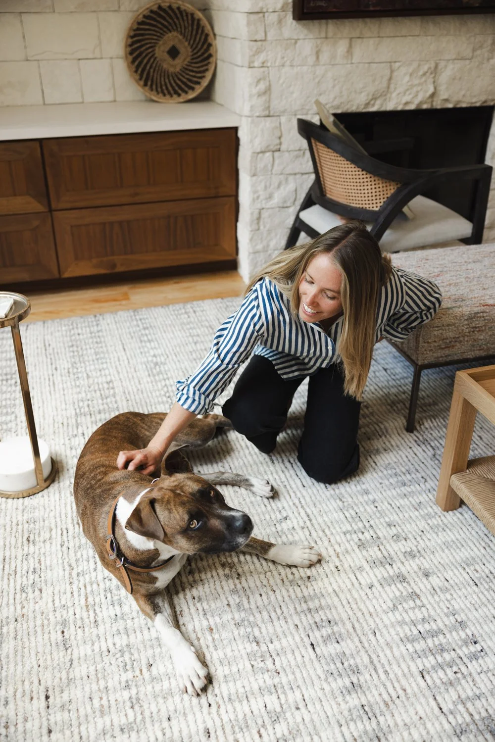 A woman with blonde hair, wearing a striped shirt, kneeling on a beige rug, petting a large brown and white dog with blue eyes, in a cozy living room with a stone fireplace, wooden furniture, and decorative items.