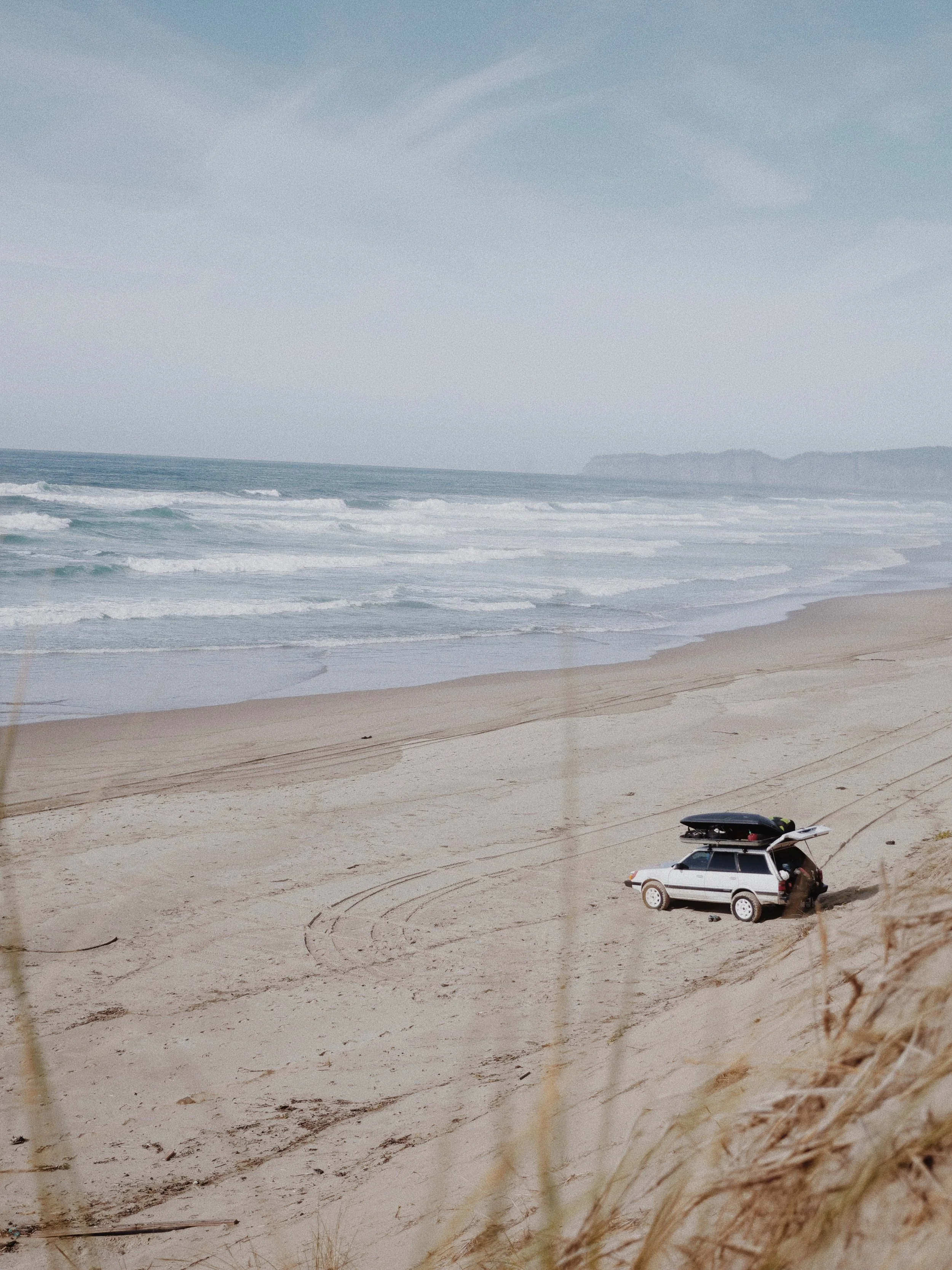 A sandy beach with ocean waves and a cloudy sky. A white car with a roof cargo box is parked on the sand in the foreground.