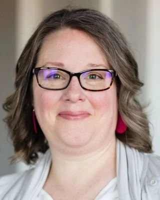 Close-up of a woman with shoulder-length brown hair, glasses, and pink earrings, smiling in an indoor setting.
