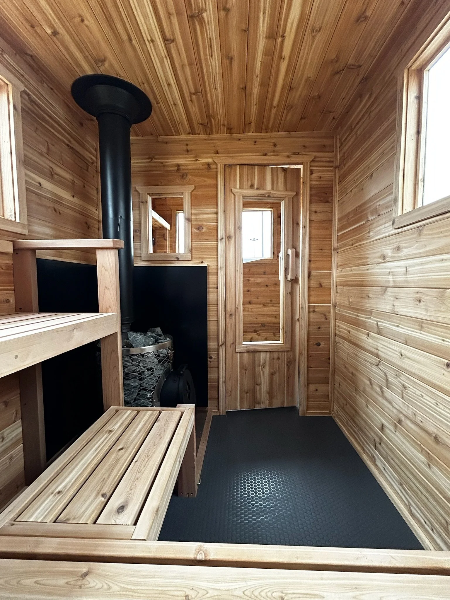 Interior of a wooden sauna with benches, a wood stove and rocks, and small windows.