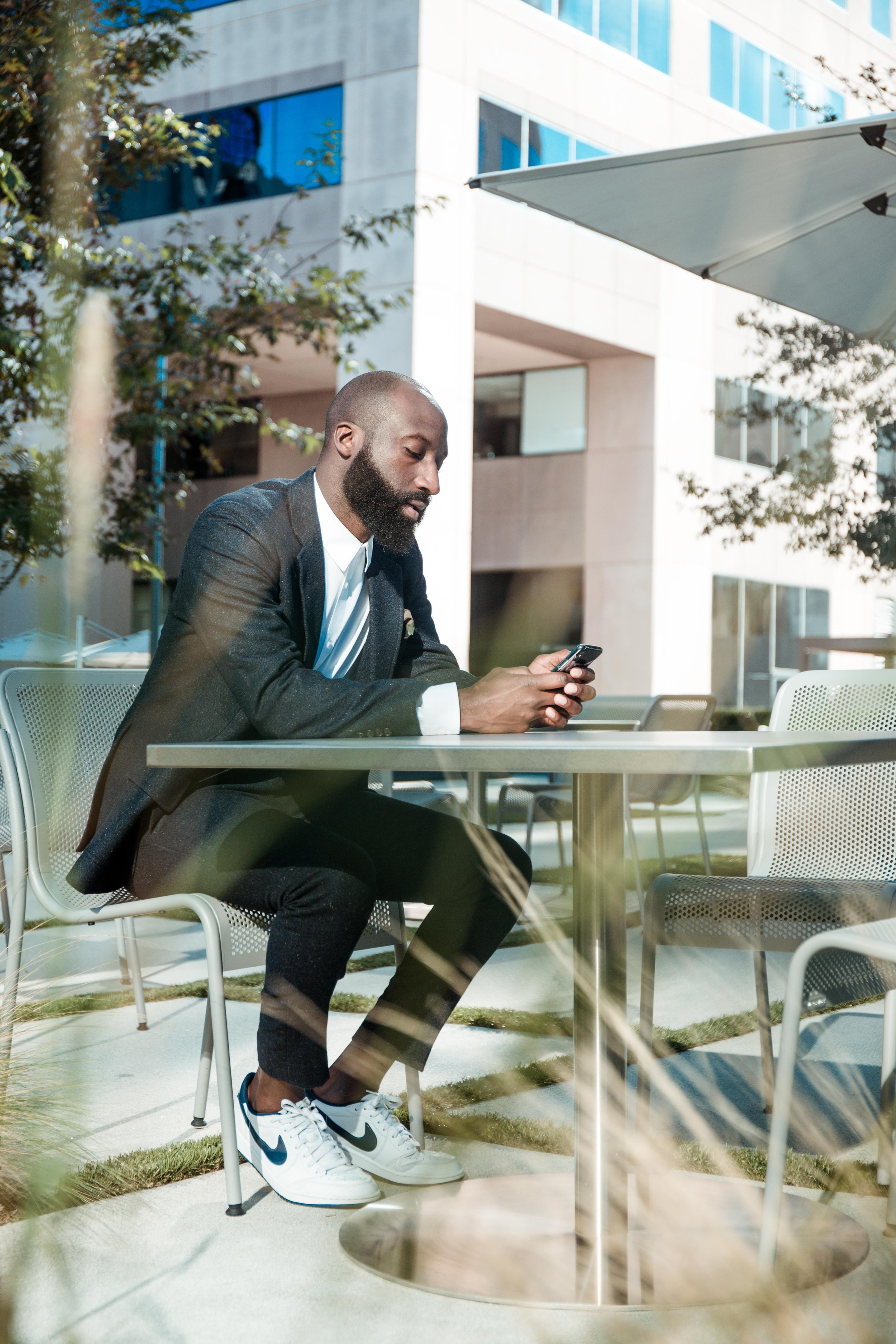 Man in a suit sitting at an outdoor table using a smartphone, with a modern building and trees in the background.