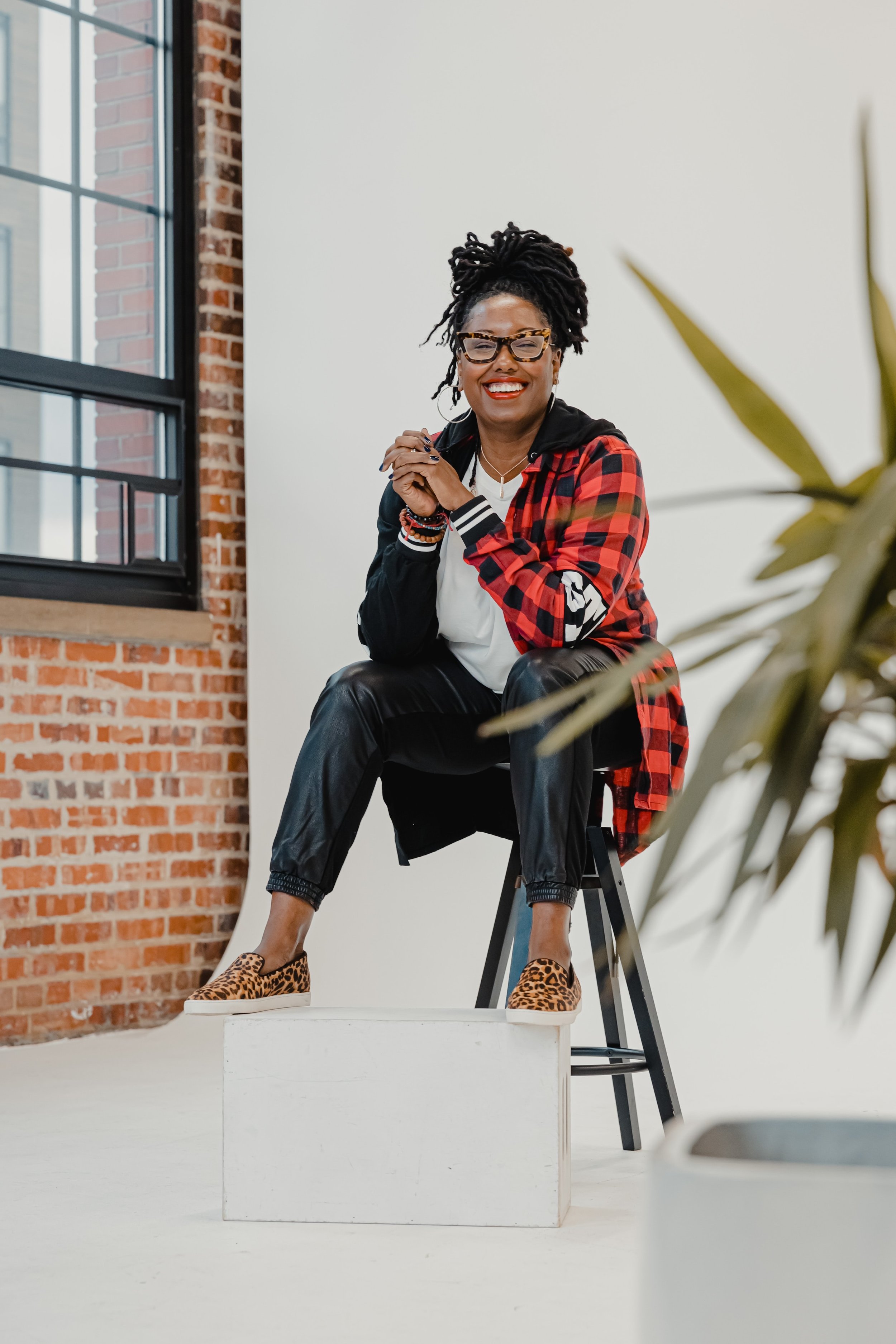 Person sitting on a stool, wearing a red plaid shirt, black pants, and leopard print shoes, smiling in a bright room with a brick wall and window.