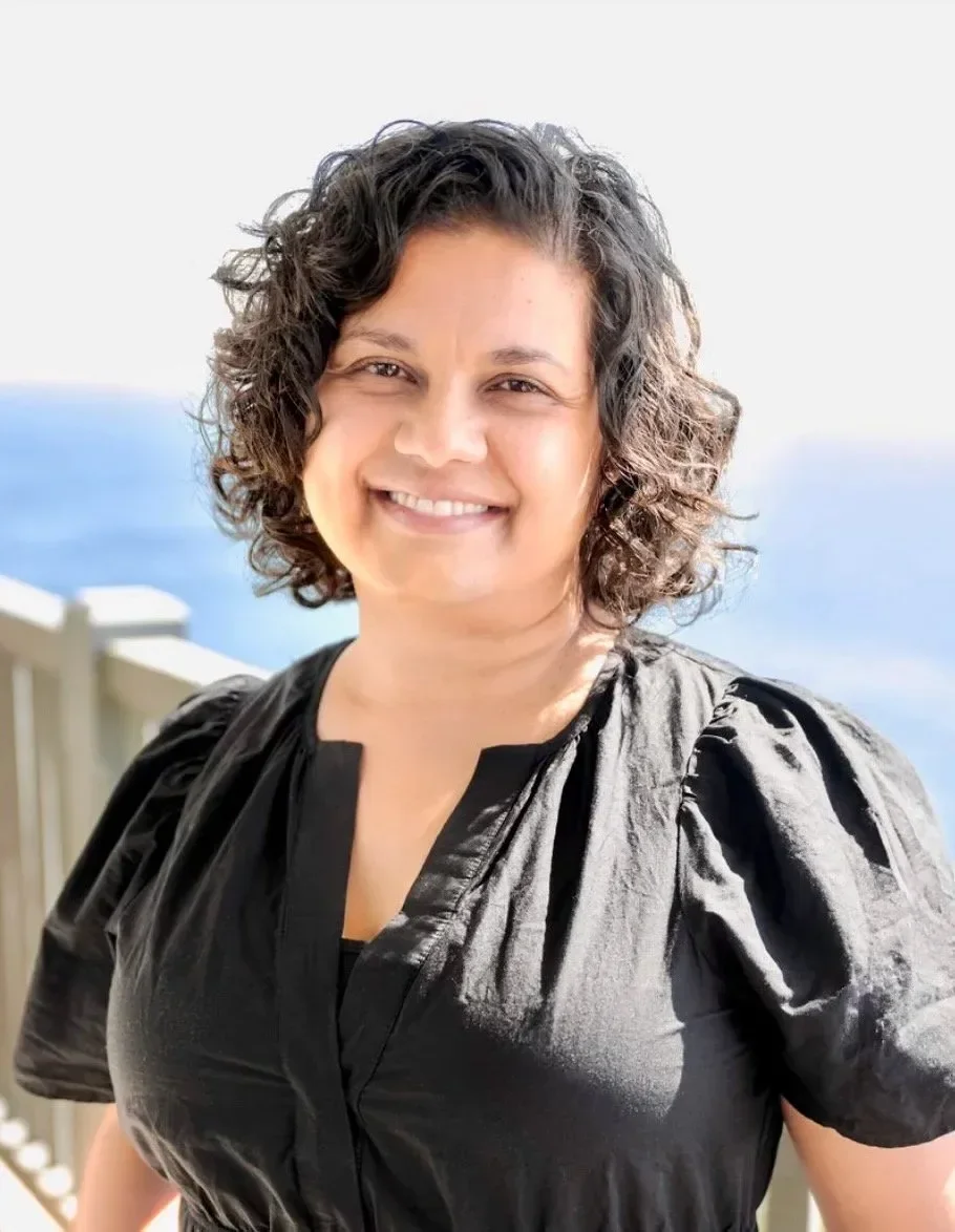 A woman with curly dark hair smiling outdoors with the ocean and sky in the background.