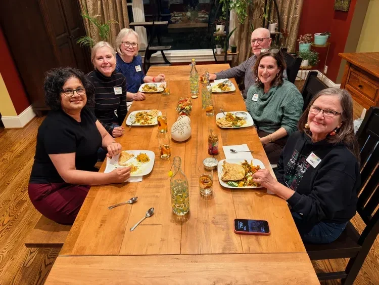 Six women sitting around a dinner table, enjoying a meal together in a warmly decorated room with wood furniture, potted plants, and curtains.