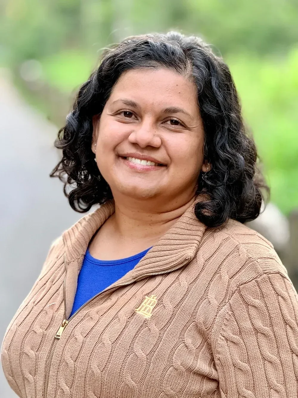 Smiling woman with dark curly hair, wearing a beige cable-knit jacket and blue shirt outdoors.