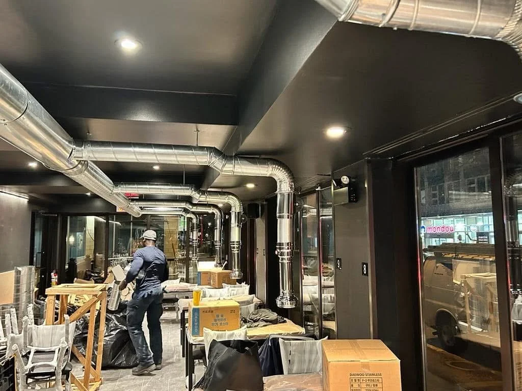 Intérieur d'un local industriel en rénovation avec des conduits exposés au plafond, des cartons empilés, et une personne travaillant.