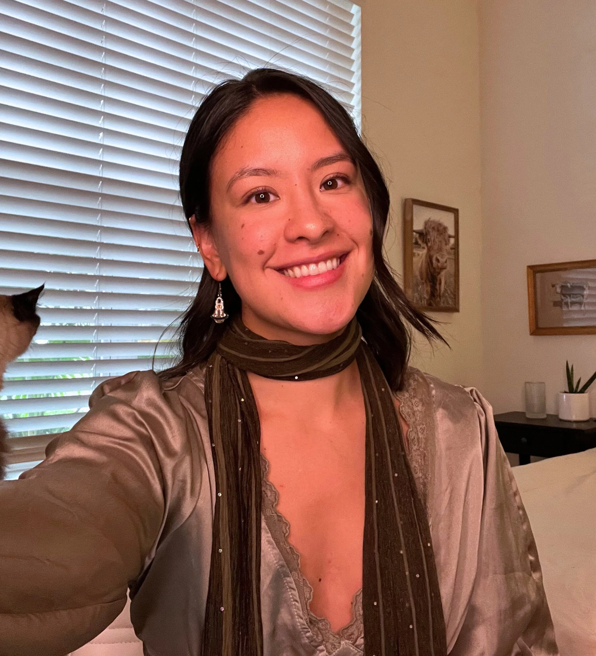 A person with dark hair wearing a black turtleneck and gold hoop earrings, standing in front of a decorative gold frame and beige draped fabric.