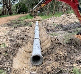 Construction site with a long concrete drainage pipe being installed in a trench, surrounded by dirt and trees.