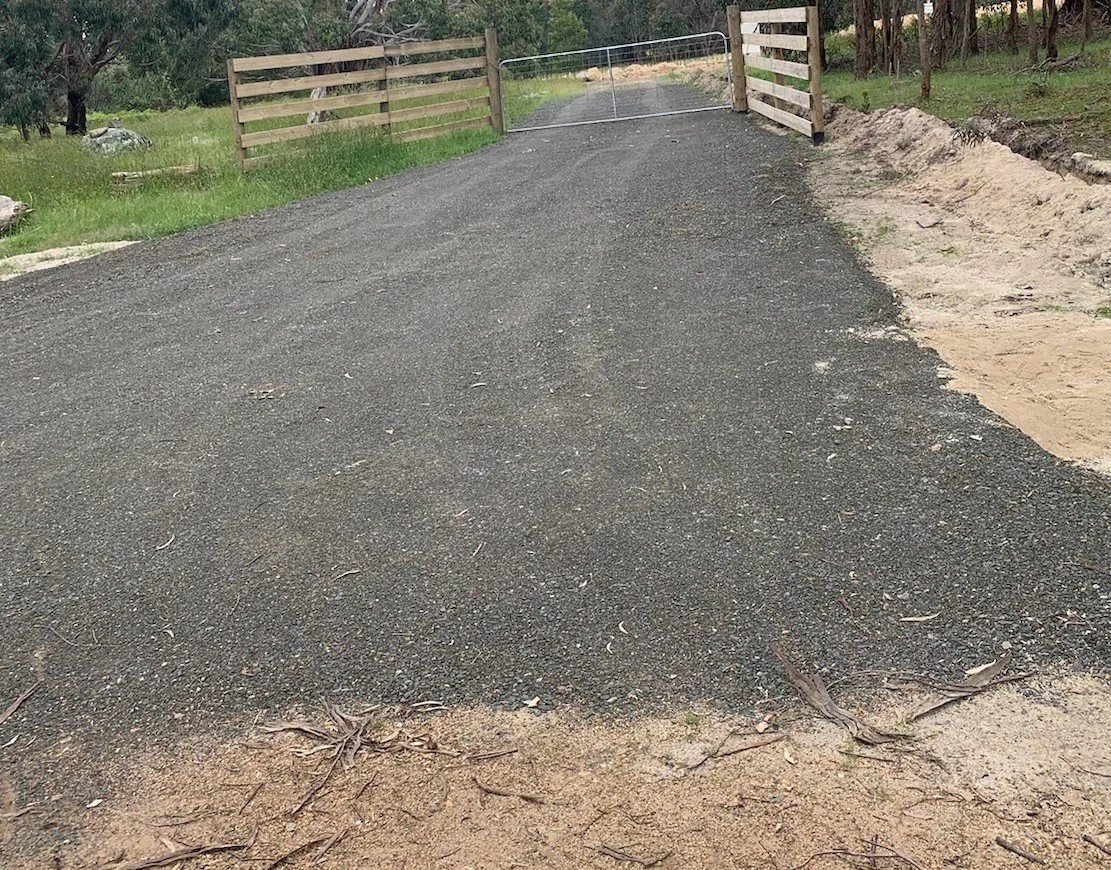 Finished concrete pipe culvert with gravel driveway leading to wooden fence and steel gate.