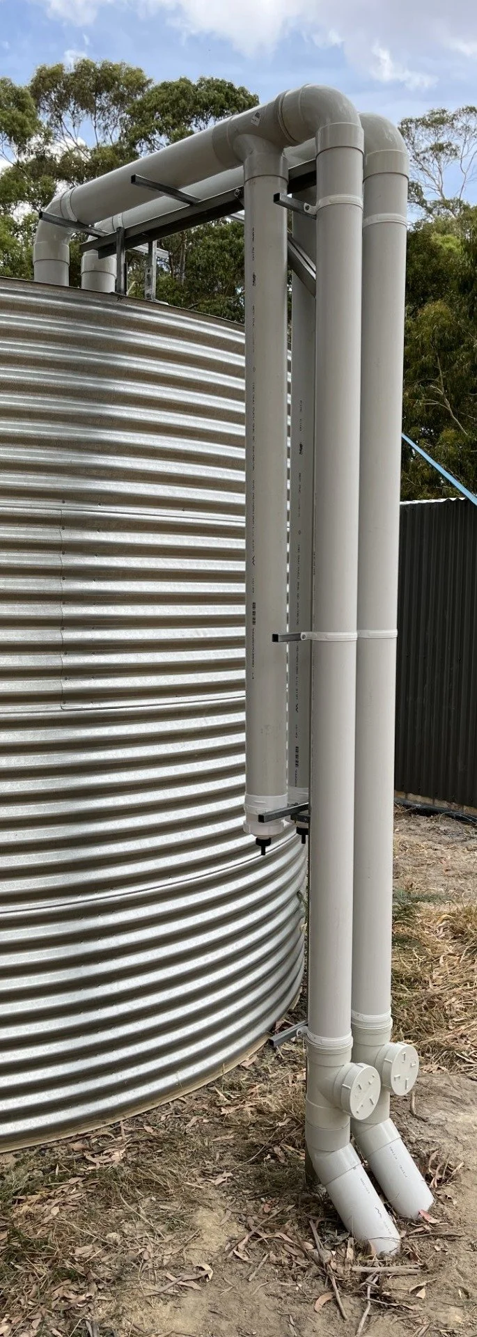 Roof rainwater collecting PVC pipes with first flush diverters supported by steel structure feeding outdoor corrugated rainwater tank.