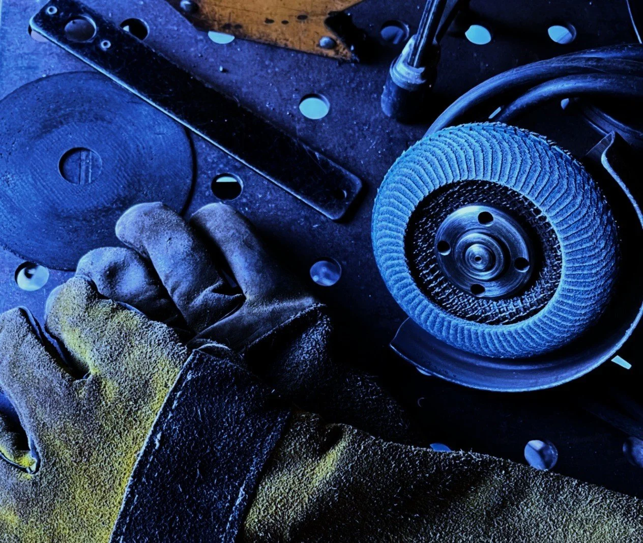 Close-up of welding gloves and angle grinding tool laying on a steel welding table illuminated with blue light.