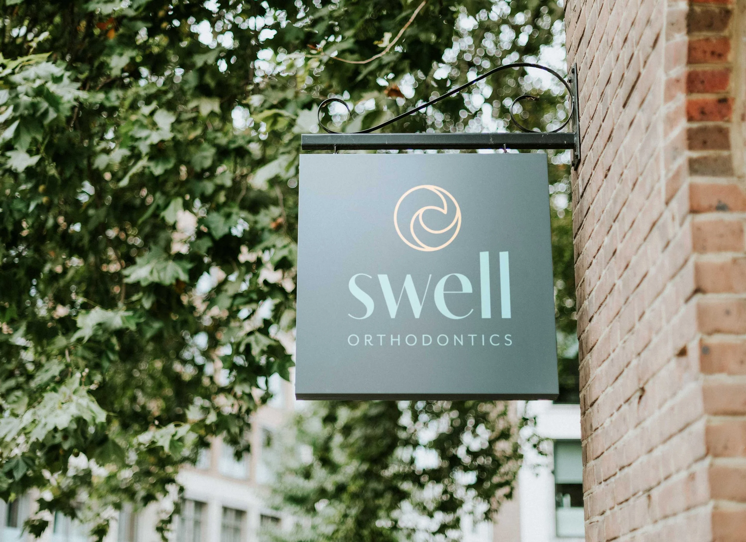 Sign for Swell Orthodontics hanging outside on a metal bracket attached to a brick wall, with green foliage in the background.