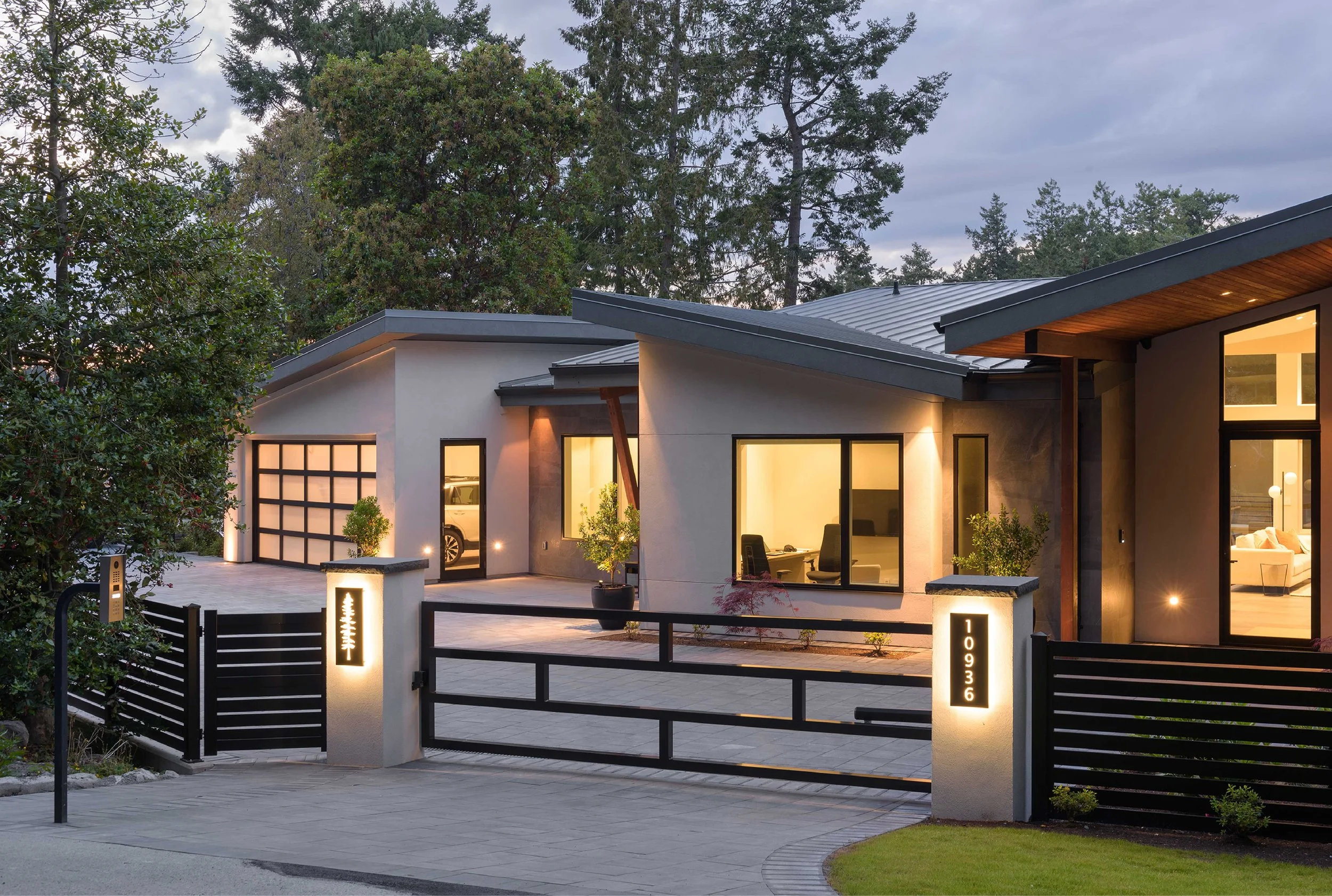 Modern house with a driveway, view of large glass windows, a garage, trees in the background, exterior lights, and a black gate with house number 10936.