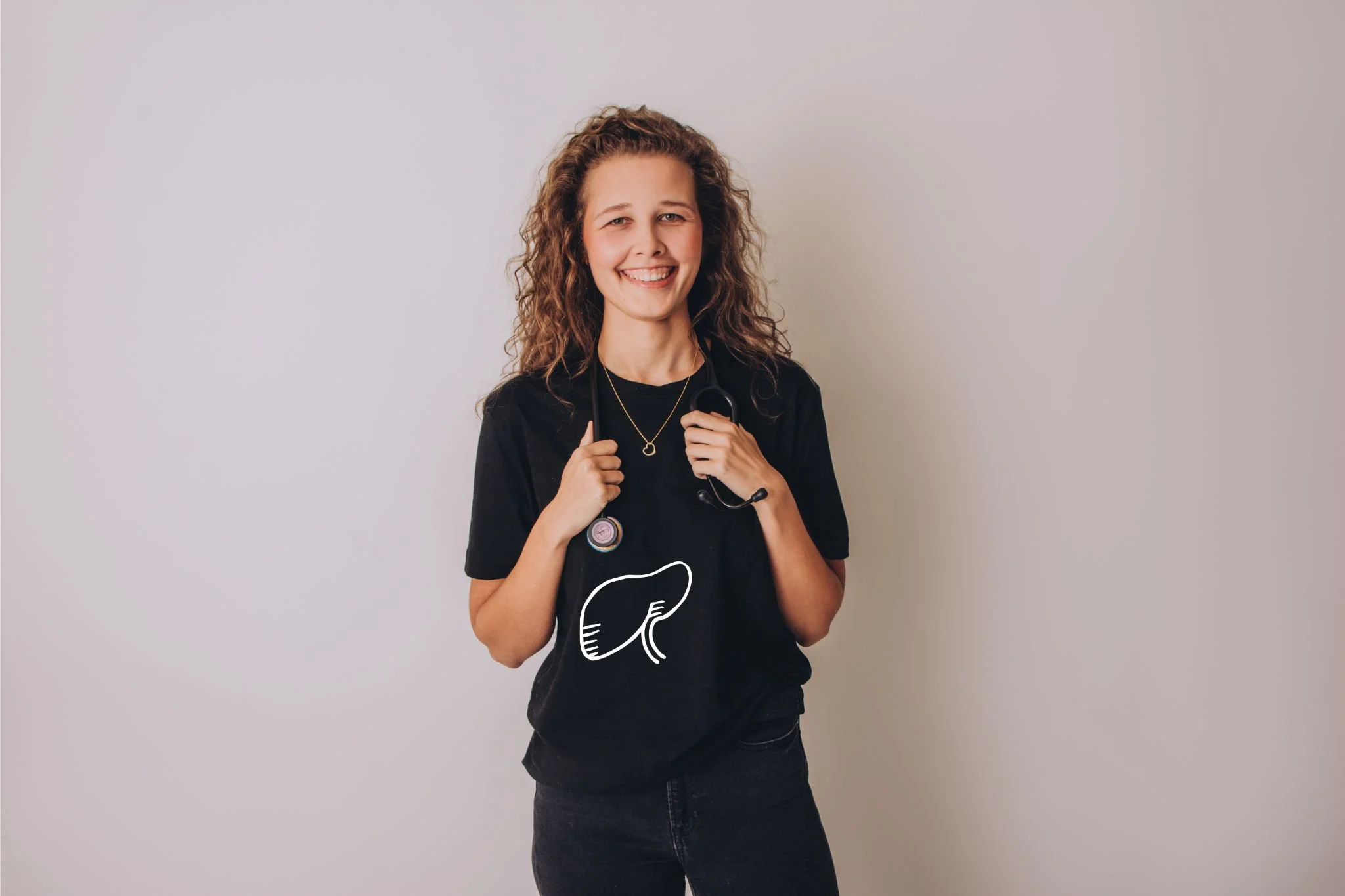 Young woman with curly hair smiling, holding a stethoscope around her neck, wearing a black t-shirt with a white liver illustration.