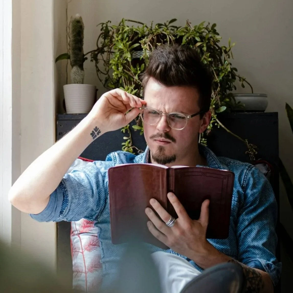 Young man seated indoors reading a leather-bound book, adjusting glasses, surrounded by plants, representing integrated faith and life in faith-based counseling.