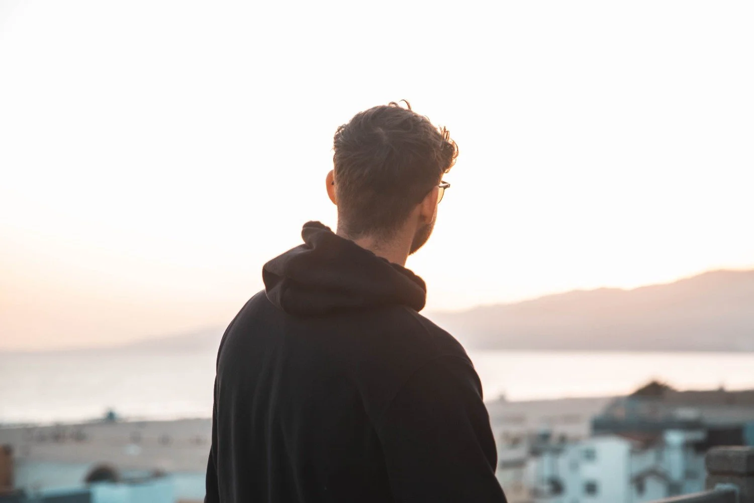 Young man with glasses facing away from camera toward a sunset horizon, representing faith-based counseling for men who want a therapist who shares their belief.