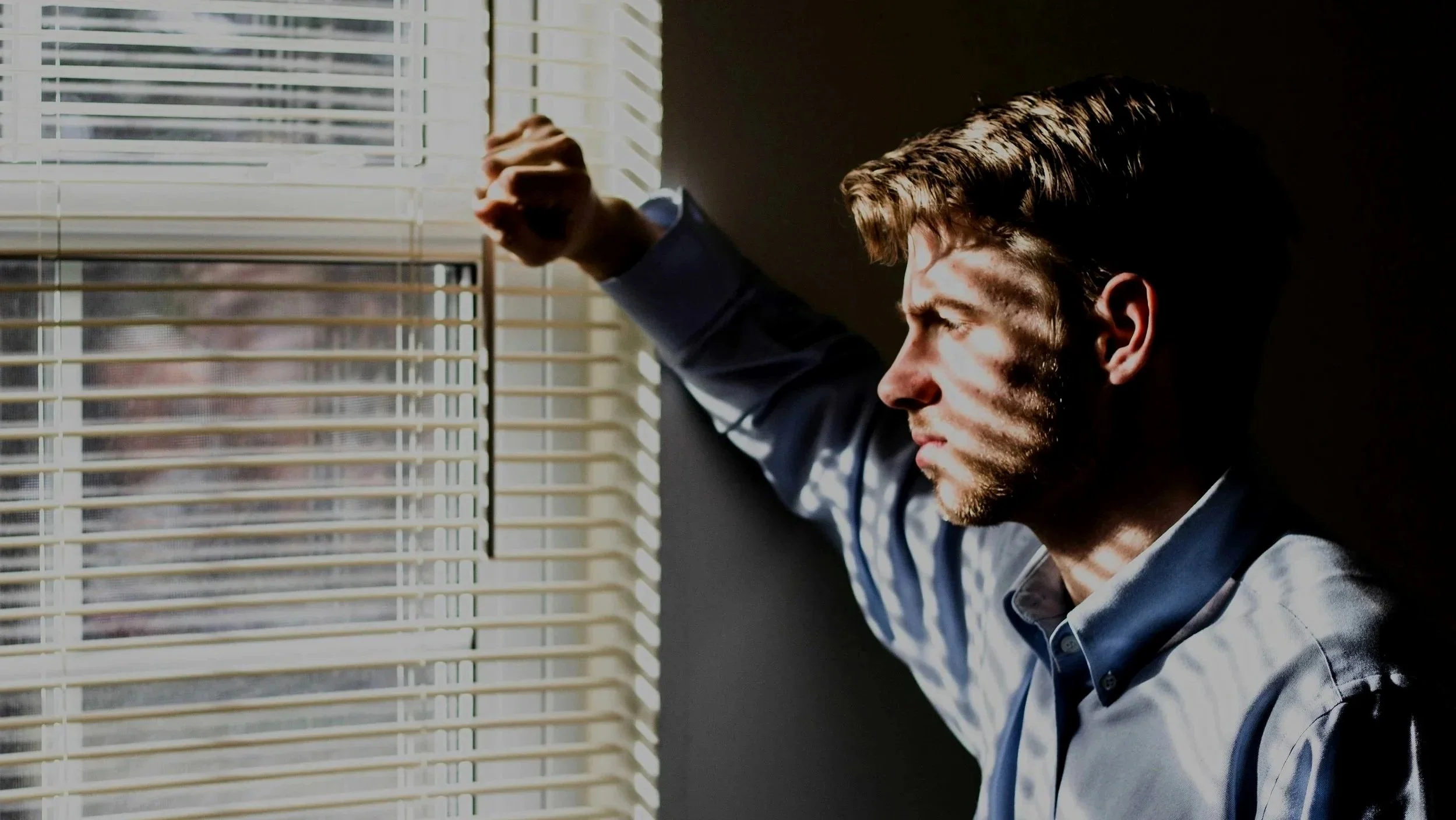 Young man in a button-down shirt standing at window blinds, representing men's anxiety counseling in Mooresville, NC.