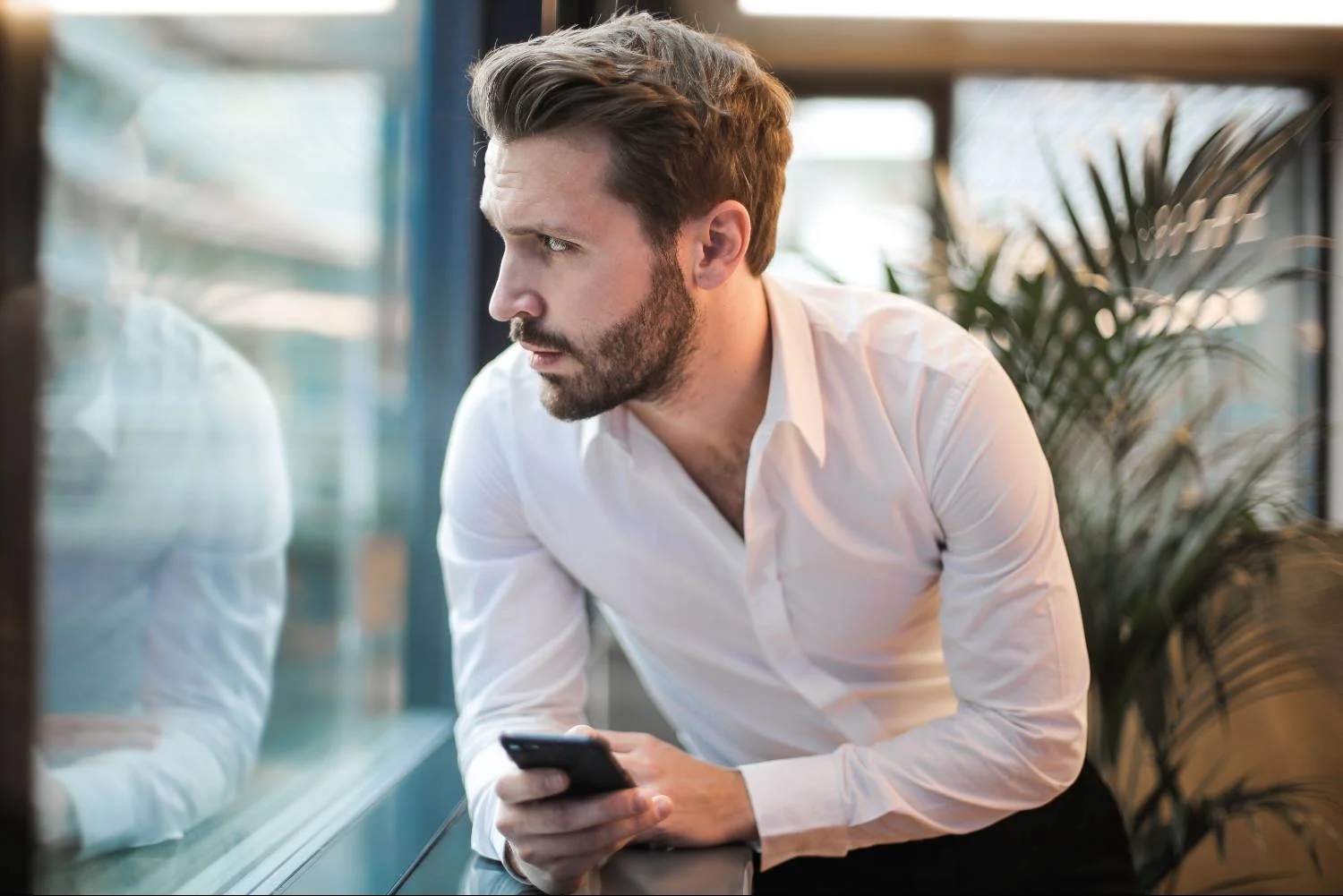 Man in white shirt leaning on windowsill holding phone, looking away, avoidant attachment pattern in men.