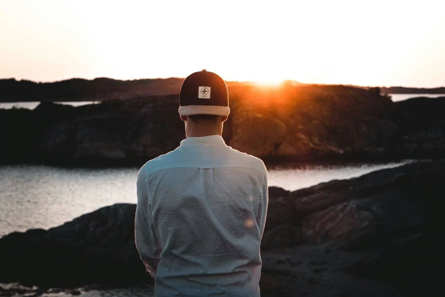 Man in baseball cap and button-down shirt standing with back to camera at rocky waterfront at sunset, representing relationship clarity from therapy for toxic relationships.