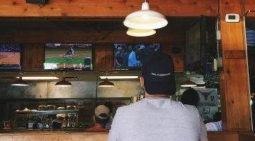 Man seen from behind wearing a cap, sitting at a bar watching a baseball game on TV.