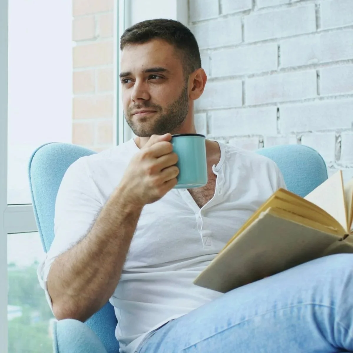 Man sitting in armchair holding mug and open book, representing faith-based counseling for men without religious shame or internal conflict.