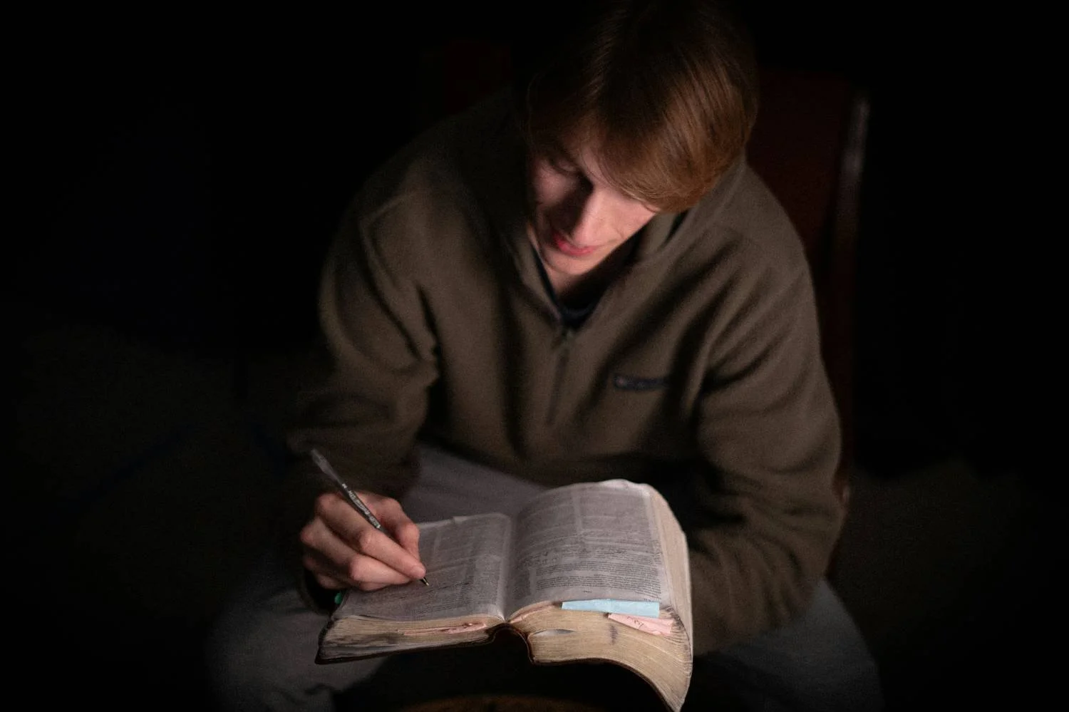 Young man writing in a heavily annotated Bible in a darkened room, illustrating scrupulosity OCD and religious anxiety counseling in North Carolina.
