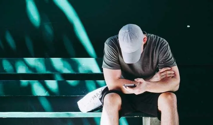 Man in a cap sitting alone on green bleachers, head down, looking at his phone with a water bottle beside him.