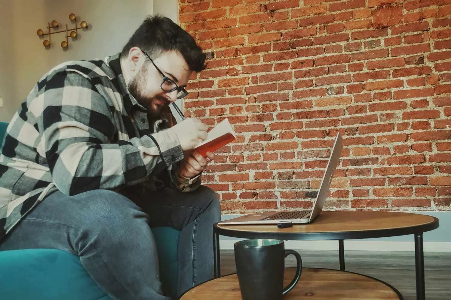 Bearded man in flannel reading a small book beside a laptop on a coffee table, representing compulsive reassurance-seeking in scrupulosity OCD treatment.