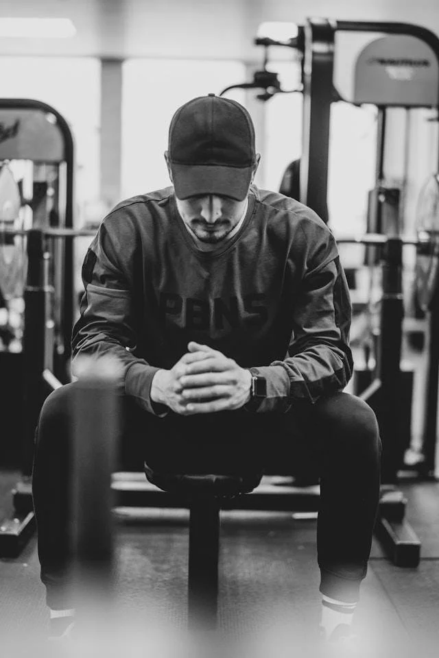 Man sitting on a gym bench, head down, hands clasped, representing the discipline men lean on when it's easier than confronting what's underneath.