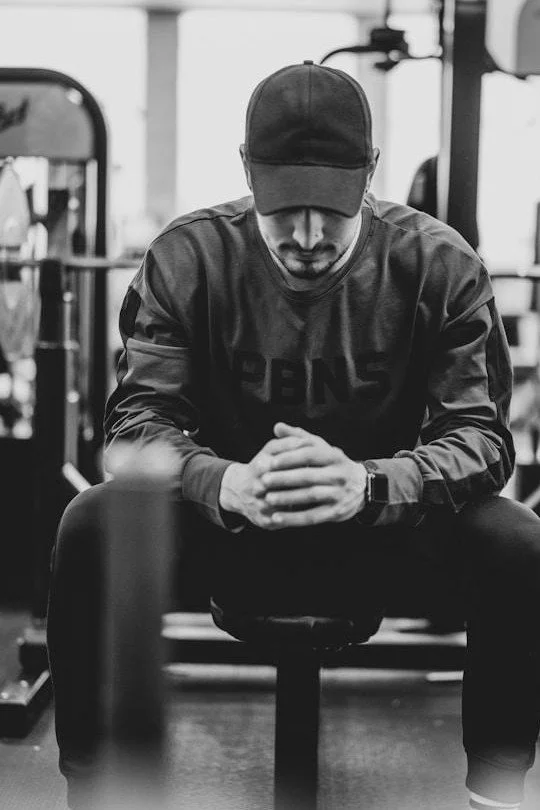 Man in baseball cap seated on gym bench, hands clasped, head down, counseling for men in Mooresville NC.