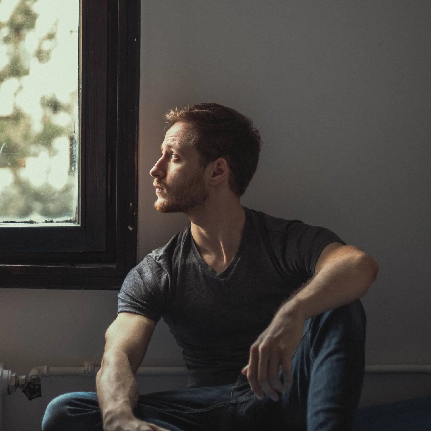 Man in gray t-shirt seated indoors looking out a window, representing avoidant attachment therapy for men in Mooresville, NC.