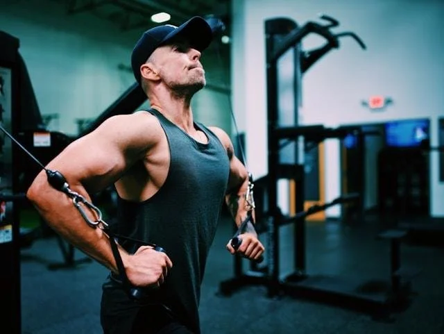 Man in a tank top and cap using a cable machine at the gym, mid-exercise.