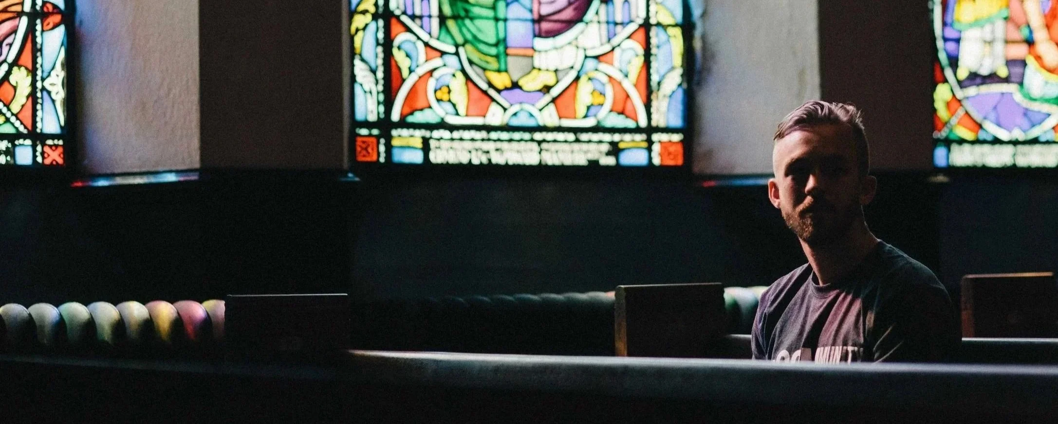 A man with a beard sits in a dimly lit church or chapel, with colorful stained glass windows behind him. The man is partially in shadow, and the stained glass windows feature intricate designs with various colors.