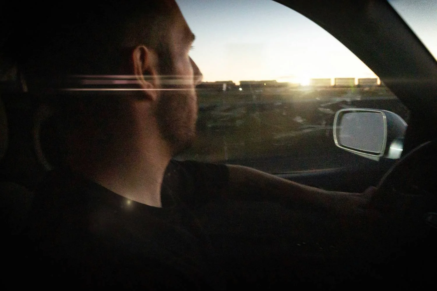Bearded man driving alone at sunset, side profile, representing a man uncertain whether to stay or leave a toxic relationship.