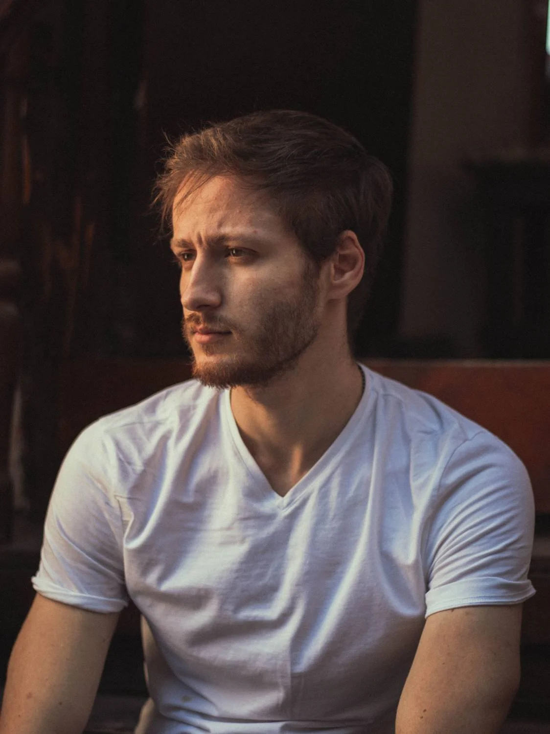 Young man in white t-shirt seated indoors looking away, representing hidden anxiety and scrupulosity in faith-based counseling for men.