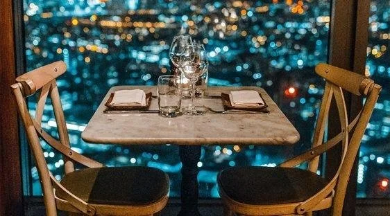A small marble table set for two by a window overlooking a lit-up city skyline at night.