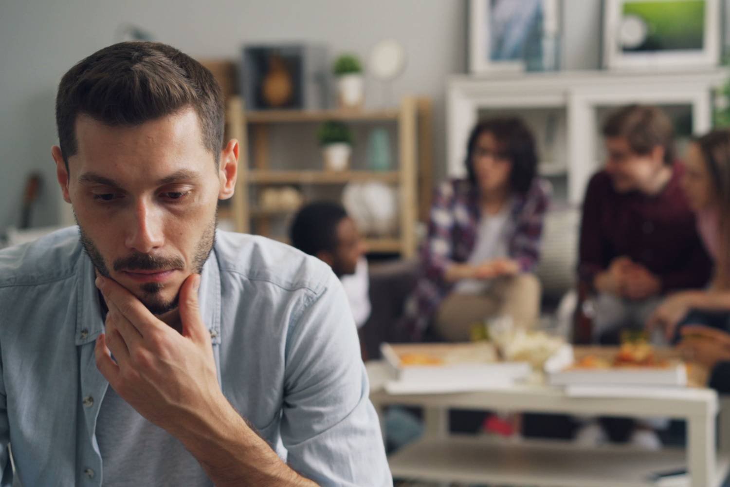Young man sitting apart from a group socializing behind him, chin resting on hand, anxious ambivalent attachment therapy for men in Mooresville NC.