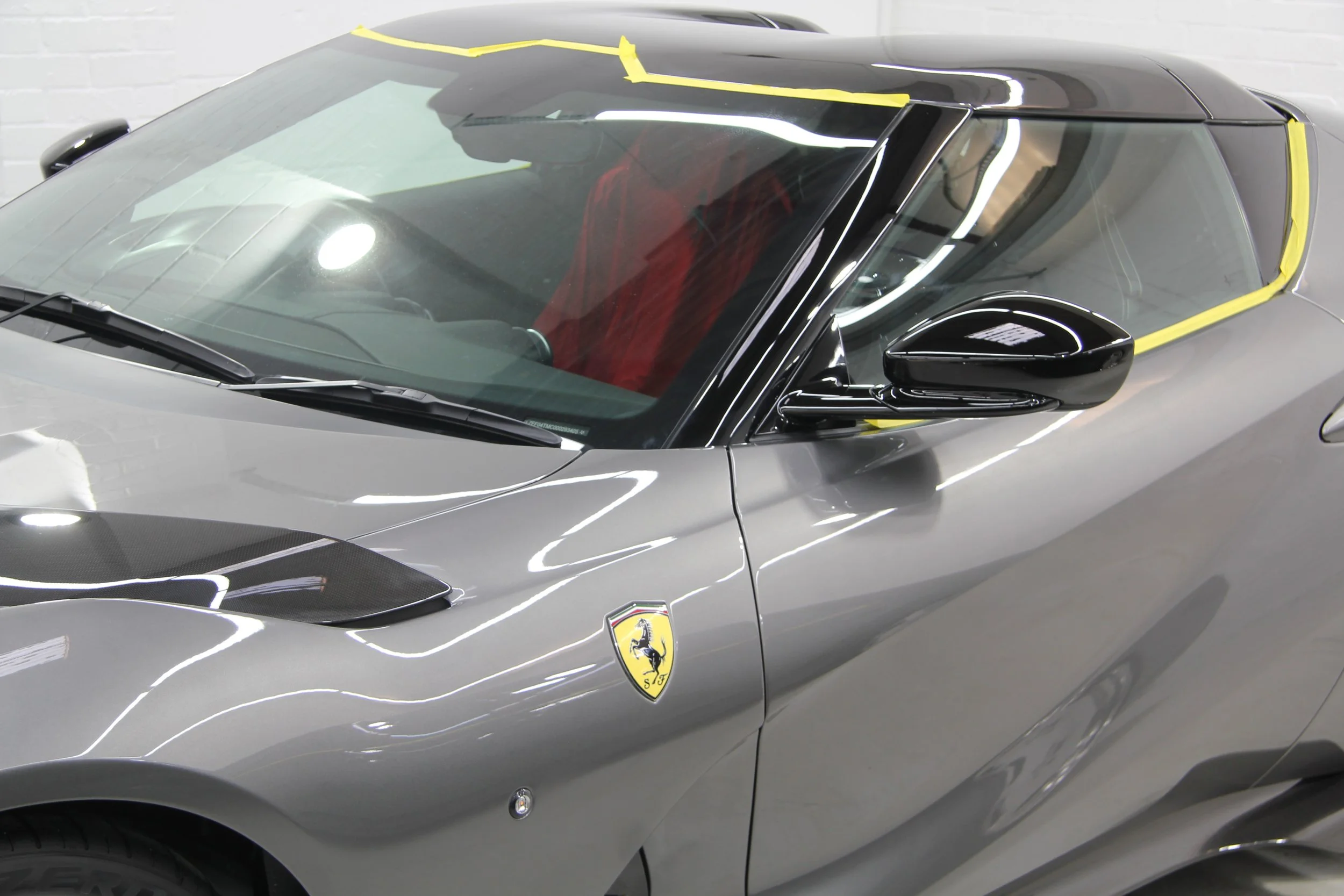 Close-up of a gray Ferrari sports car with yellow masking tape on windshield and door, featuring the Ferrari logo and black side mirror.