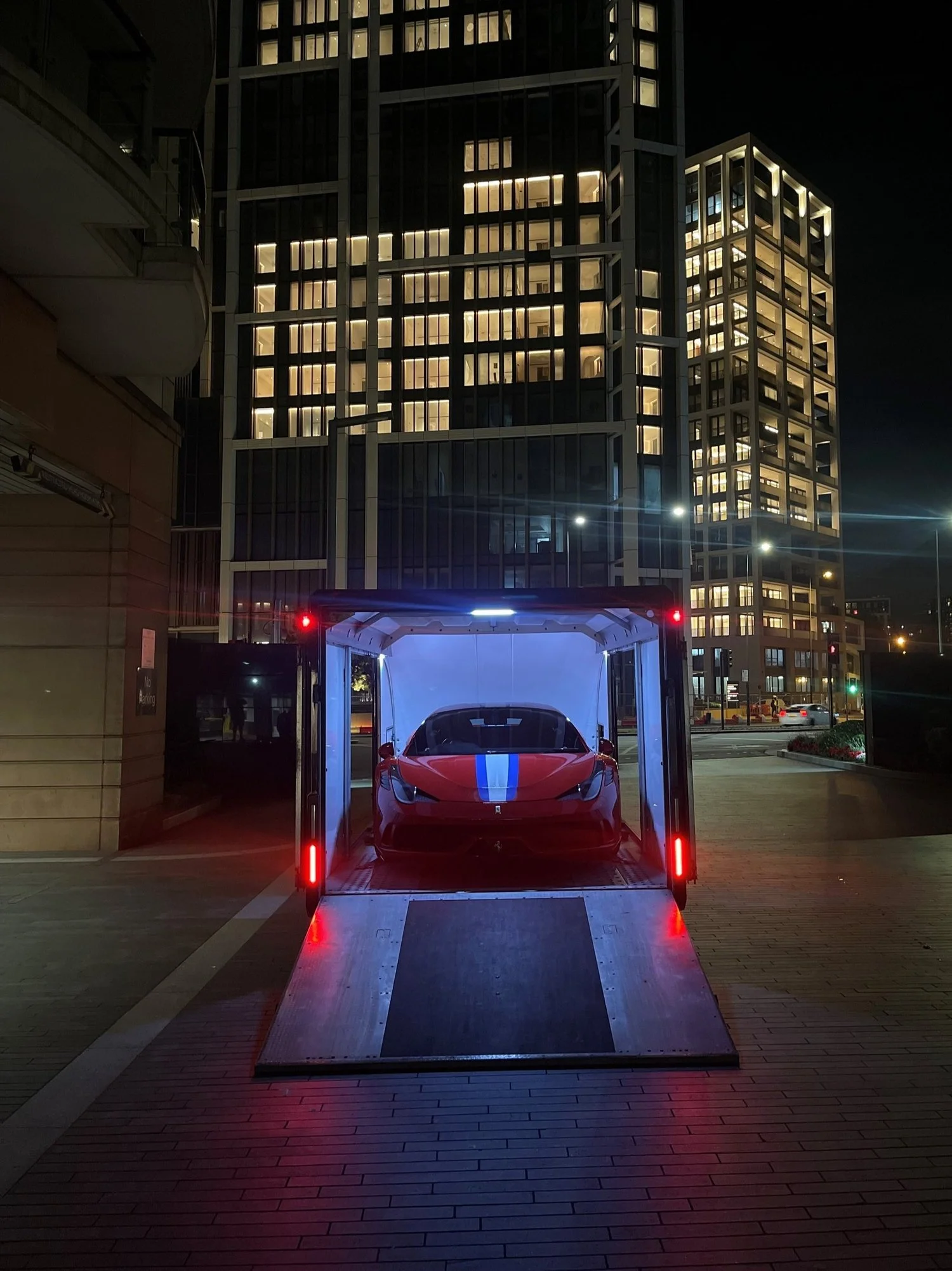 Red sports car with blue stripe inside a trailer at night, city buildings in background.