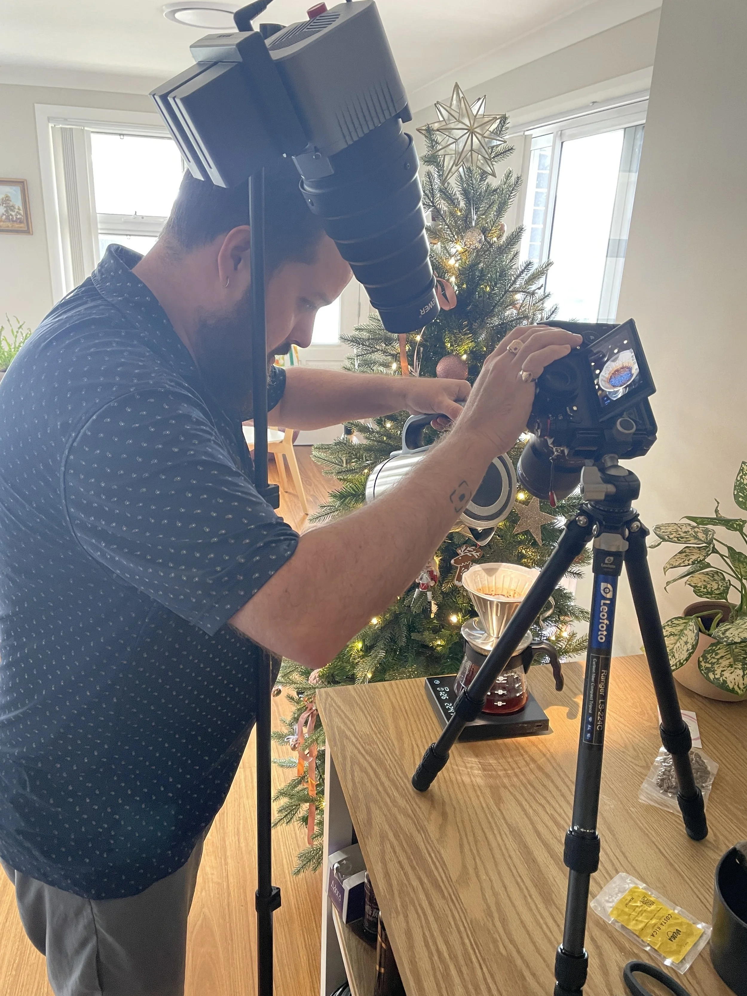 A man with a beard and earrings making coffee using a Chemex coffee maker on a wooden table with a Christmas tree decorated with ornaments and a star on top in the background.