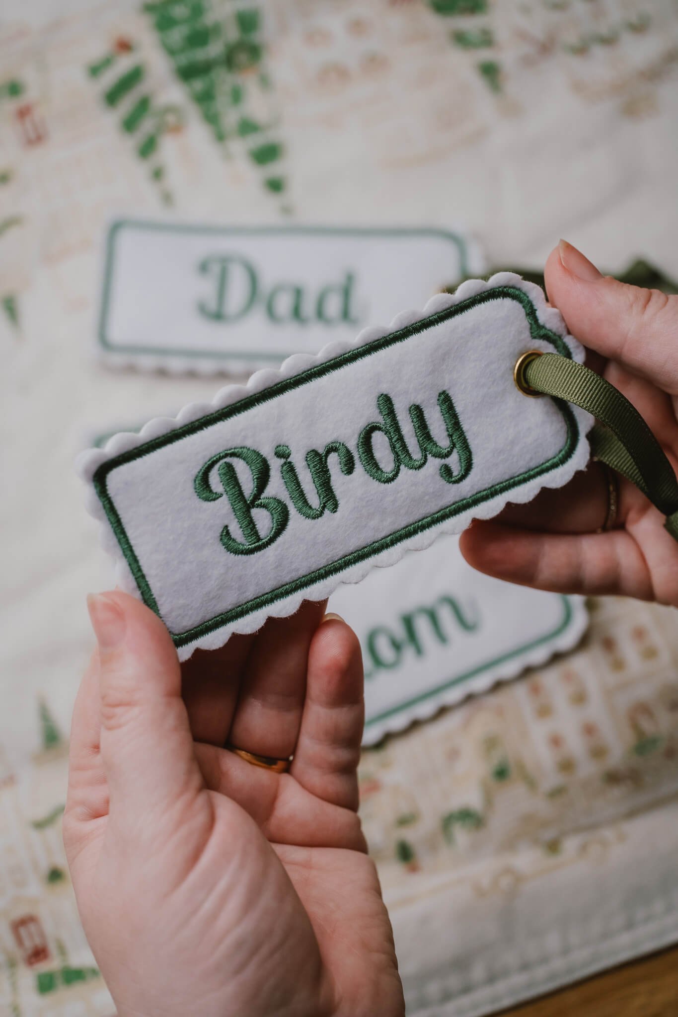 Close-up of a person's hands holding a fabric name tag that says "Buddy" with a background of similar name tags that say "Dad" on a patterned surface.