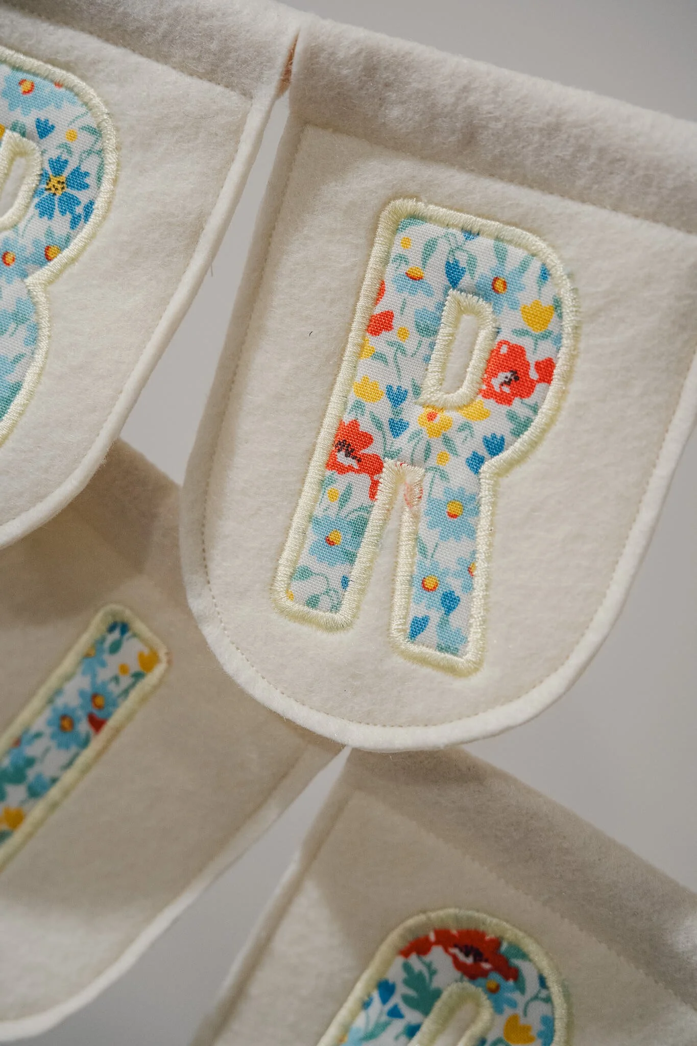 Close-up of fabric letters with floral pattern embroidery, beige felt backing, and rounded edges.