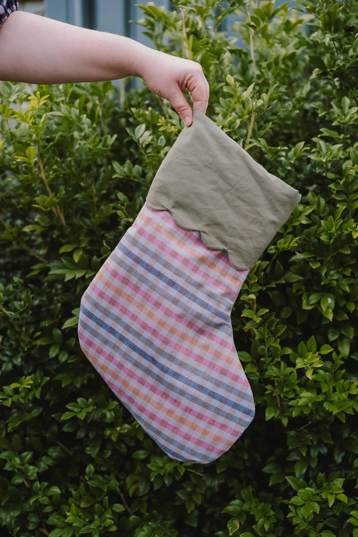 A person's hand reaches into a green bush while standing outside. They are holding a checkered cloth with pink, orange, blue, and white colors.