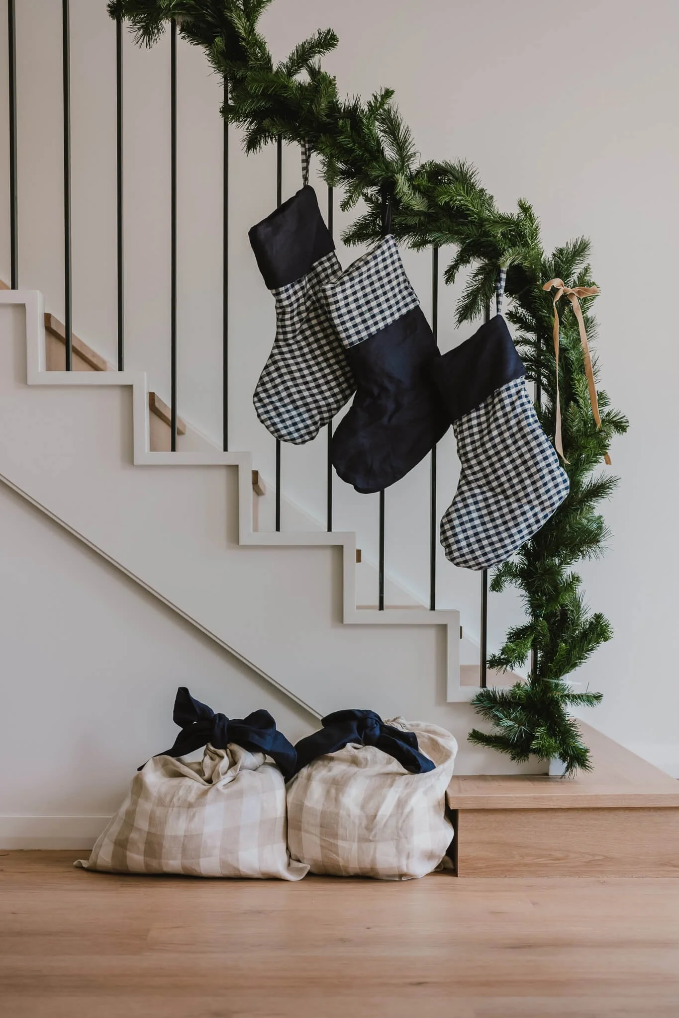 Decorated staircase with a green garland and three Christmas stockings hanging from the garland. There are two large laundry bags on the floor at the bottom of the staircase.