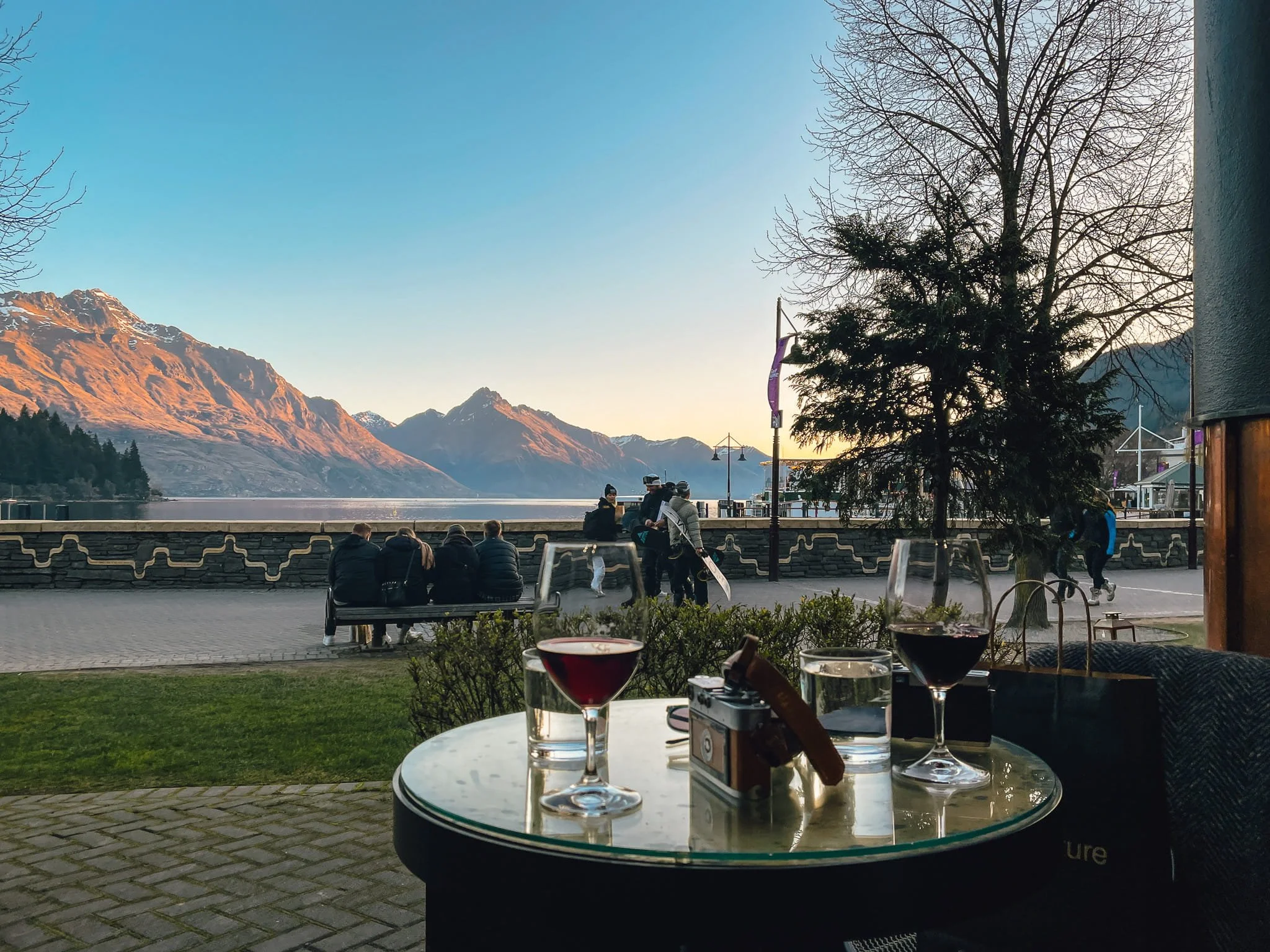 Wine and a film camera on a table overlooking Lake Wakatipu at sunset in Queenstown, with the Remarkables mountains lit in golden light