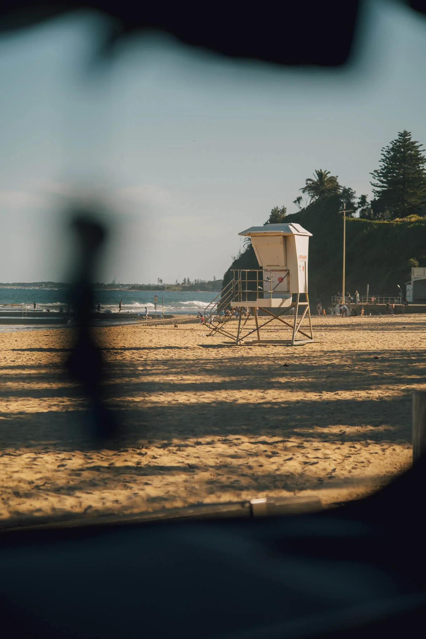 View of a sandy beach with a lifeguard tower, ocean waves, and some people in the distance, seen through a window or barrier.