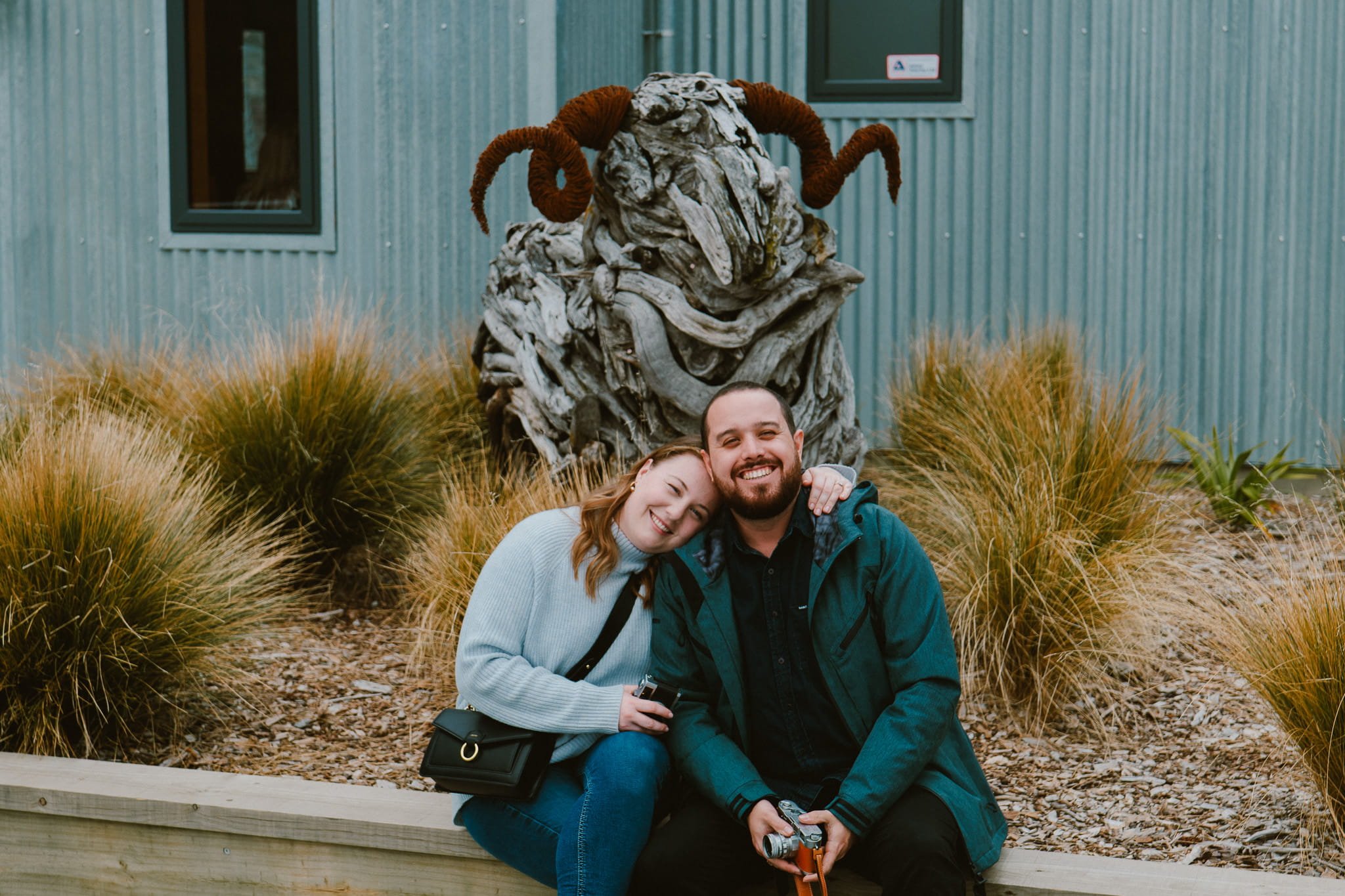 Couple sitting together outside Mt Rosa winery with a sculptural ram in the background, Gibbston Valley