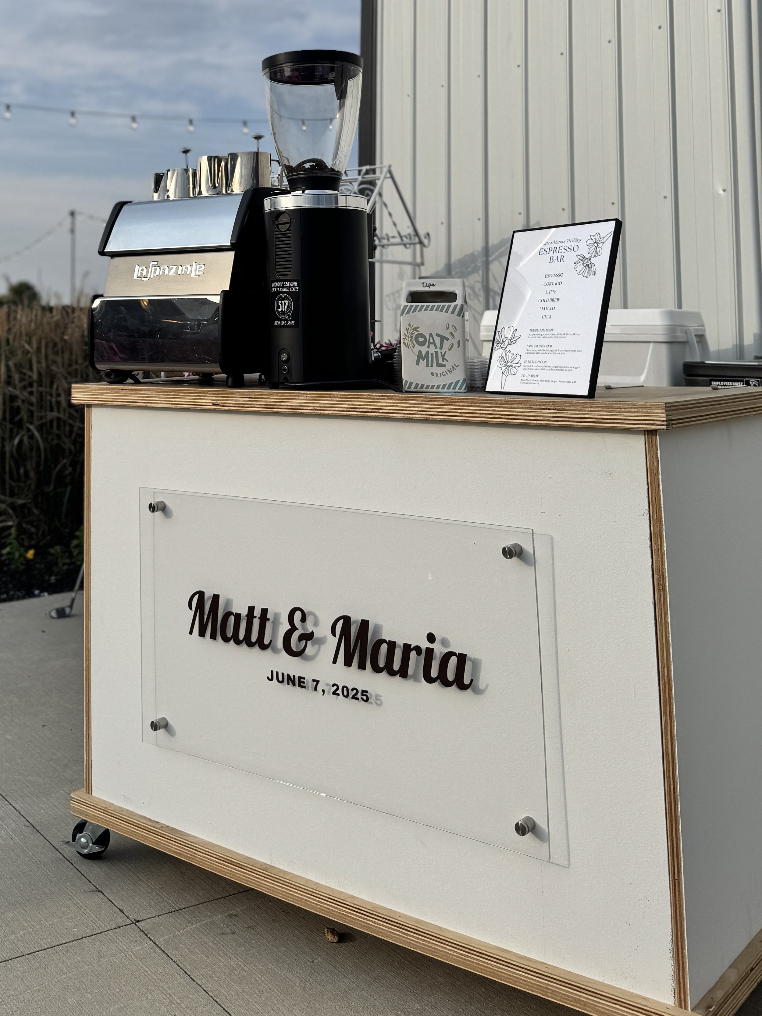 Outdoor espresso bar with coffee machine, oat milk container, and menu sign featuring a custom sign for a wedding date June 7, 2025.